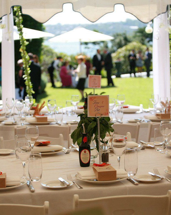 Sit down wedding in outdoor marquee with multi-lingual table number in front, guests on lawn and Sydney Harbour in background
