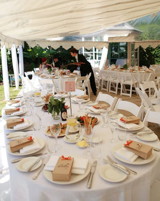 Sit-down wedding dinner in marquee being set up by female waiter as musicians set up in background